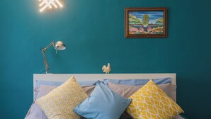 Une chambre avec des murs de couleur turquoise, un lit agrémenté d’oreillers à motifs bleus et jaunes, une lampe de chevet et un tableau encadré accroché au mur.