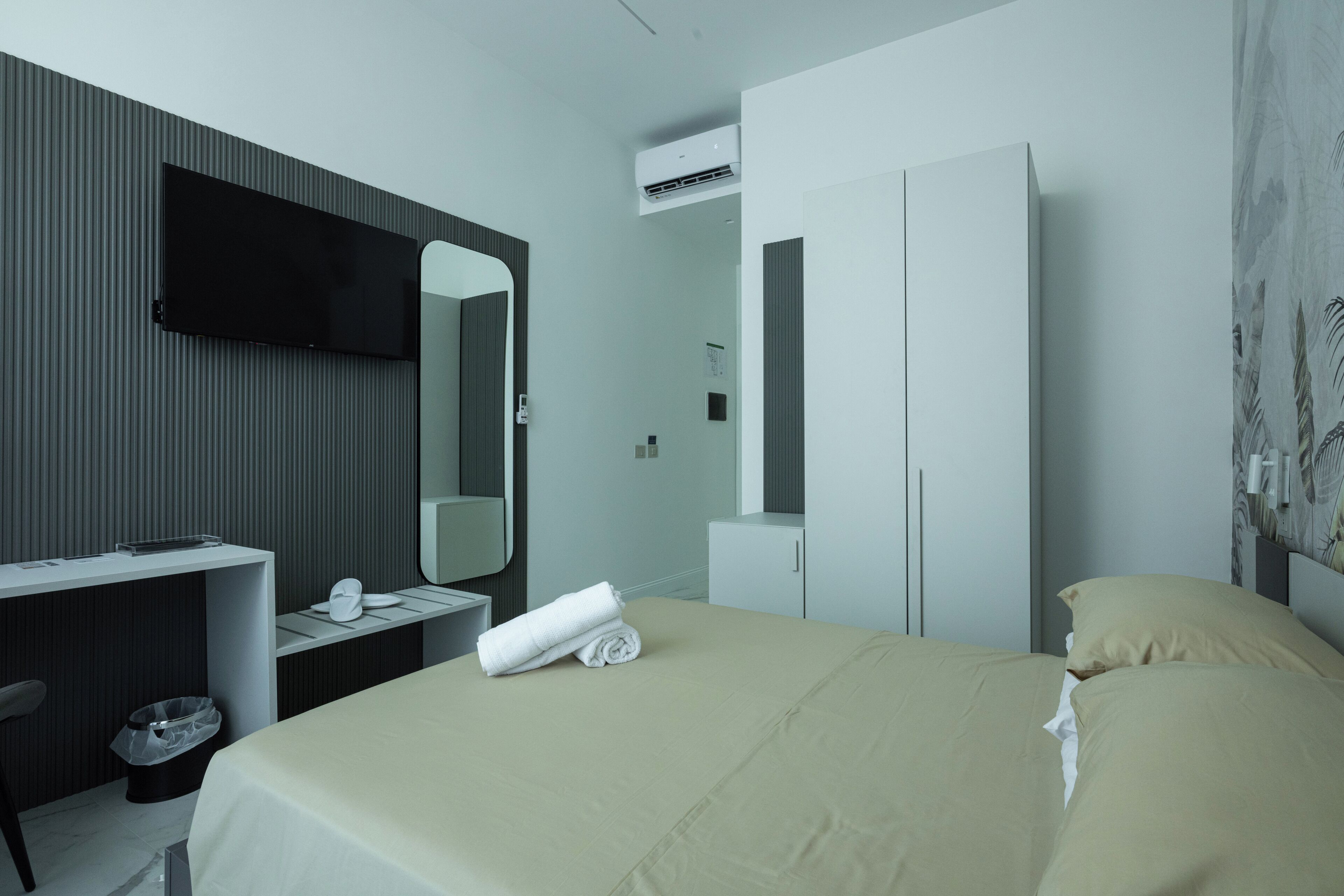 A hotel room with a bed, a desk, a chair, a TV, a wardrobe, and an air conditioning unit.