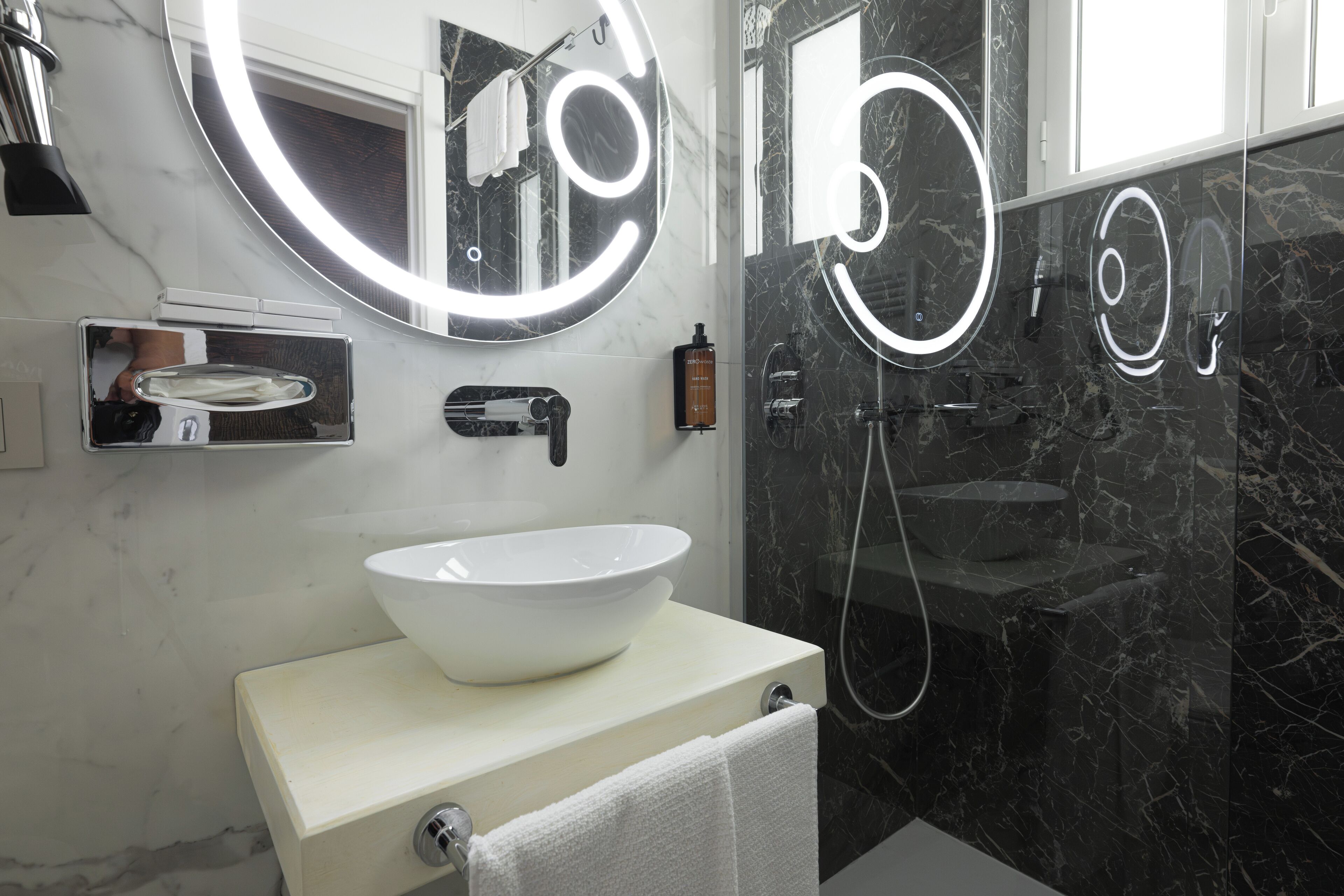 A modern bathroom with a white sink, a large mirror with circular lights, and a black marble shower area.