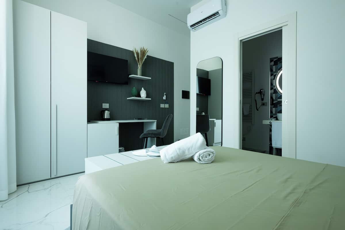 Une chambre d’hôtel moderne équipée d’un lit, d’un bureau, d’une chaise, d’un miroir et d’une salle de bain visible à travers une porte ouverte.