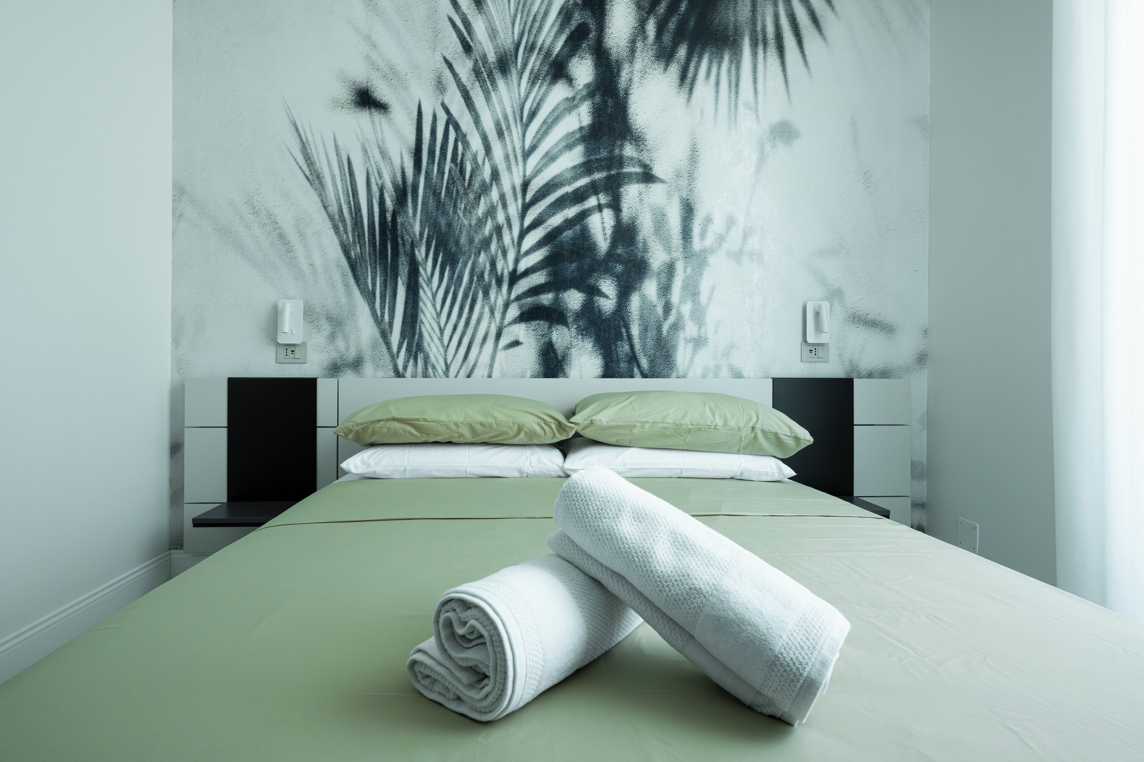 Una cama bien tendida con ropa de cama verde, dos almohadas y toallas enrolladas en una pared blanca con un mural de hojas de palmera.