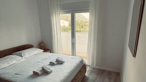 A bedroom with a bed, a nightstand, a white rug, and a view of a mountain through a window. - Cozy 2-bedroom apartment in small place Ostrvica near Omiš (Ostrvica)