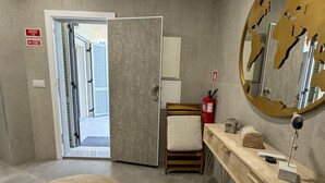 A modern bathroom with a large mirror, a wooden chair, a round rug, and a wooden vanity with a fire extinguisher.