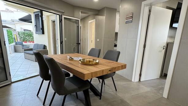 A modern dining area with a wooden table and black chairs, a sliding glass door leading to an outdoor patio, and a kitchen visible through an open door.