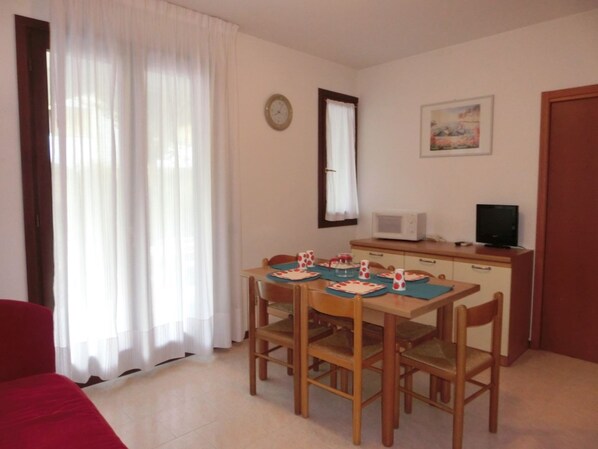 A room with a dining table, chairs, a microwave, a television, and a clock on the wall. - Apartment with garden in residence with pool (Bibione)