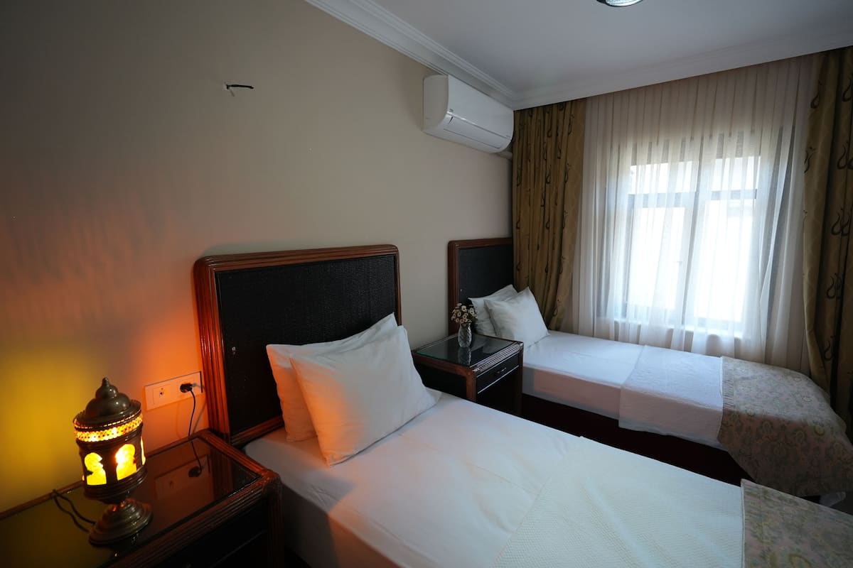 A hotel room with two beds, a nightstand with a lamp, an air conditioner, and a window with curtains.