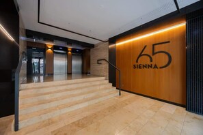 A modern hotel lobby with a staircase leading to the upper floors, a wooden wall with the number 65, and a glass elevator.