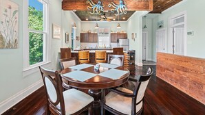 Un coin repas avec une table et des chaises en bois, un ventilateur de plafond et, au fond, une vue sur la cuisine.
