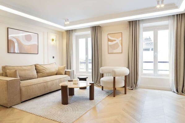A modern living room with a beige sofa, a round coffee table, and a white armchair. There are two large windows with curtains, and artwork on the walls. - A Splendid 2-BR/2BA with Services in Opéra (Paris)