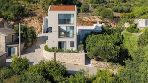 Un bâtiment moderne à deux étages, doté d’une façade en pierre, de grandes fenêtres et d’un toit recouvert de tuiles rouges, entouré d’une végétation luxuriante et d’autres bâtiments.