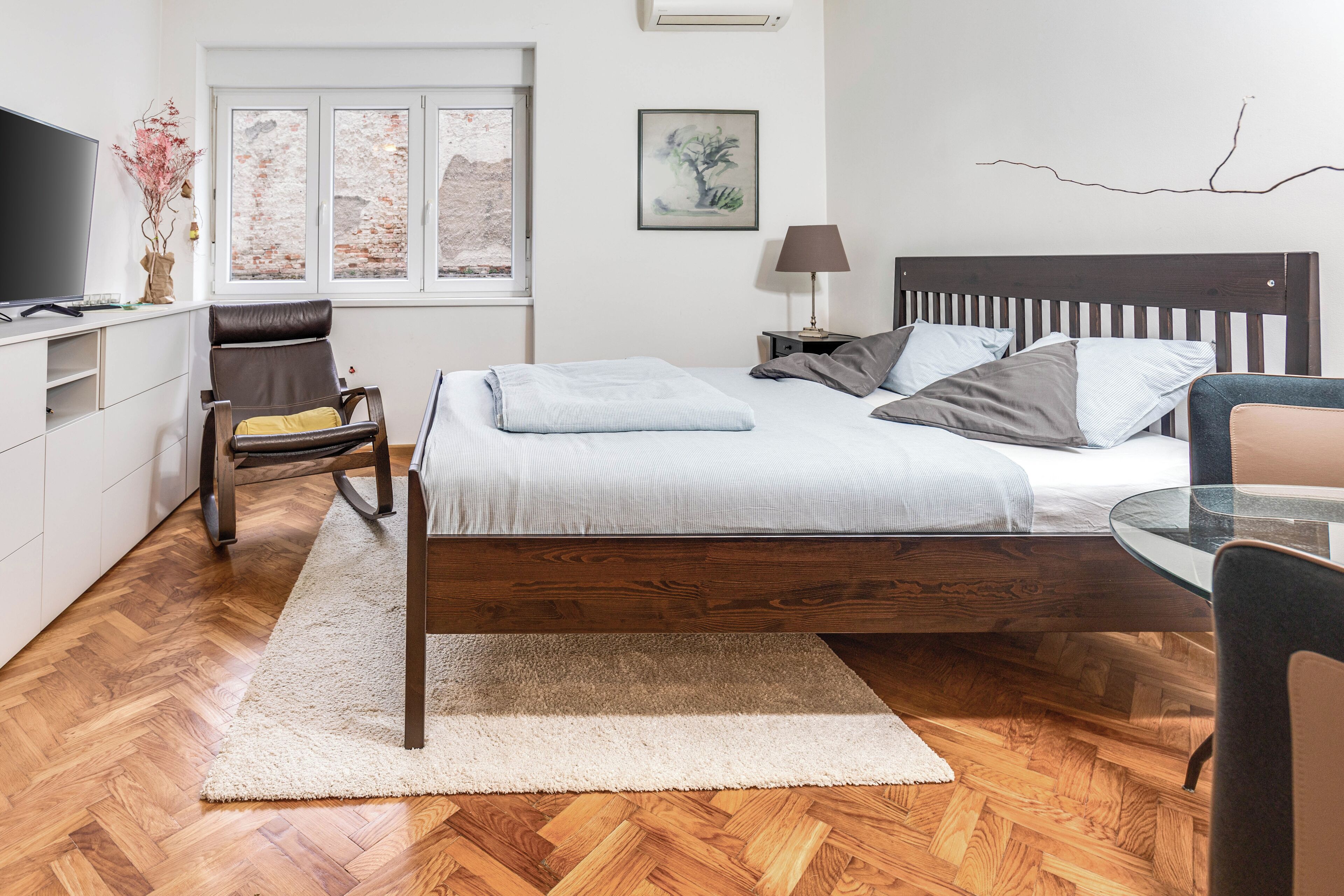 Une chambre à coucher avec un lit en bois, un bureau avec une télévision, une chaise et une fenêtre donnant sur un mur de briques.
