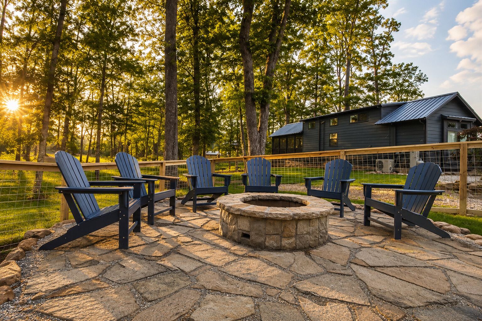 A stone patio with Adirondack chairs and a fire pit, surrounded by a wooden fence and a house with a metal roof.