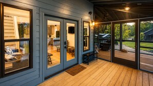 Une véranda couverte avec une terrasse en bois, un coin repas avec une table et des chaises, et une vue sur un jardin avec un barbecue et des arbres.