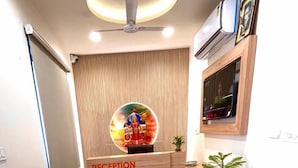 A reception area with a wooden reception desk, a cushioned sofa, a potted plant, and a ceiling fan. - Hotel Rajvi Residency A Unit of JAS Hotels & Resorts (Bikaner)