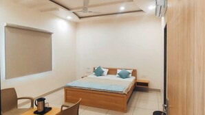 A bedroom with a wooden bed, a desk with a chair, a coffee maker, and a nightstand. - Hotel Rajvi Residency A Unit of JAS Hotels & Resorts (Bikaner)