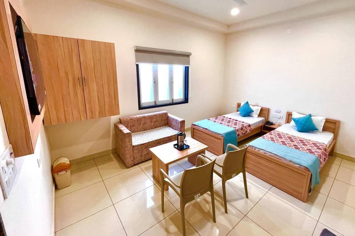 A small, well-lit room with two single beds, a sofa, a table with chairs, and a ceiling fan.