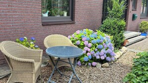 Un bâtiment en briques avec une fenêtre, deux chaises en osier, une petite table ronde et un parterre de fleurs avec des hortensias.