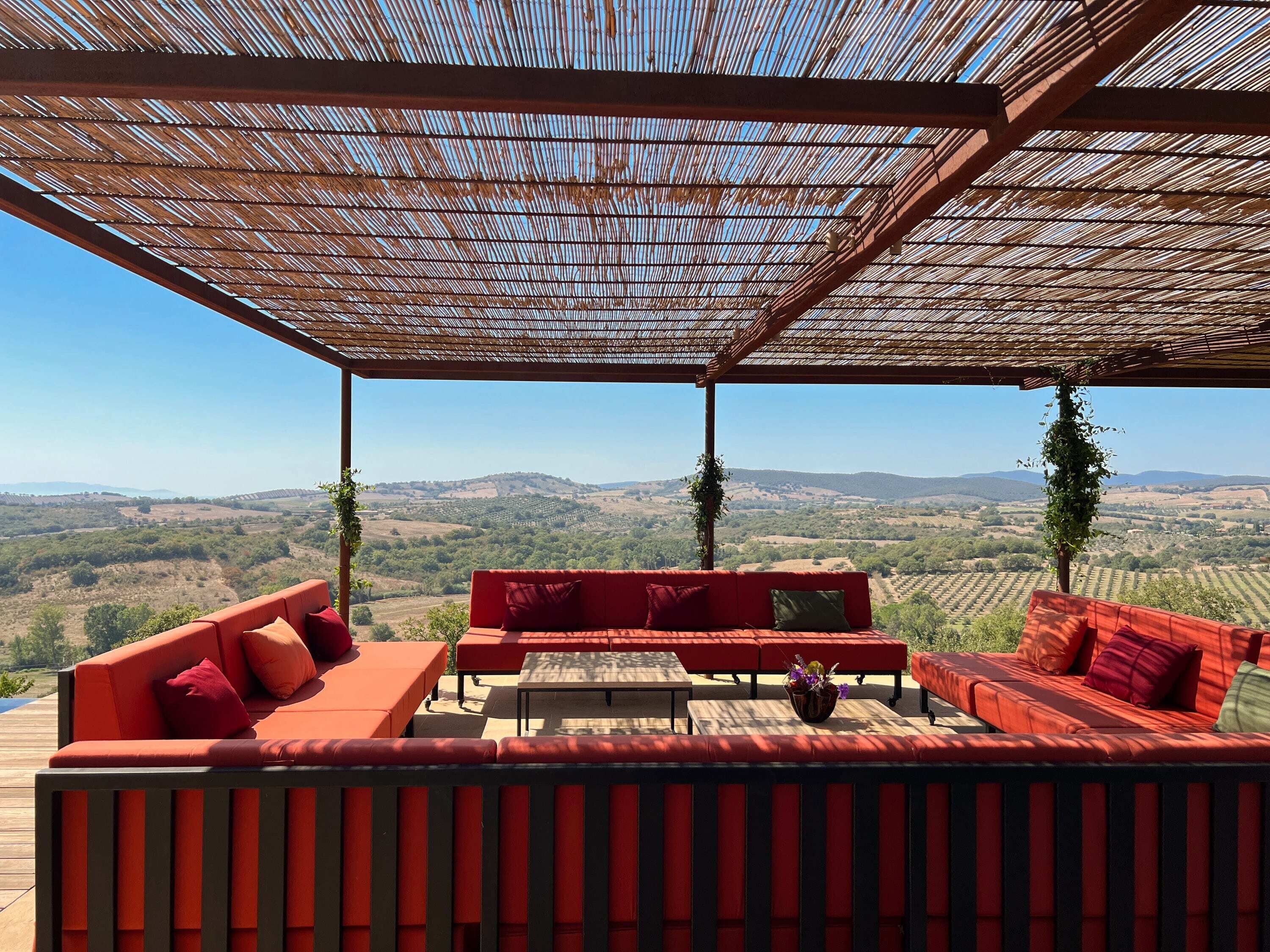 Ein überdachter Außenbereich mit roten Sofas und einem Tisch, mit Blick auf eine hügelige Landschaft.