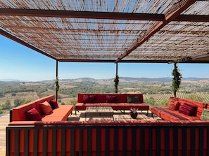 A covered outdoor seating area with red sofas and a table, overlooking a hilly landscape. - Maniero in Maremma, luxury villa  in Tuscany (Montiano)