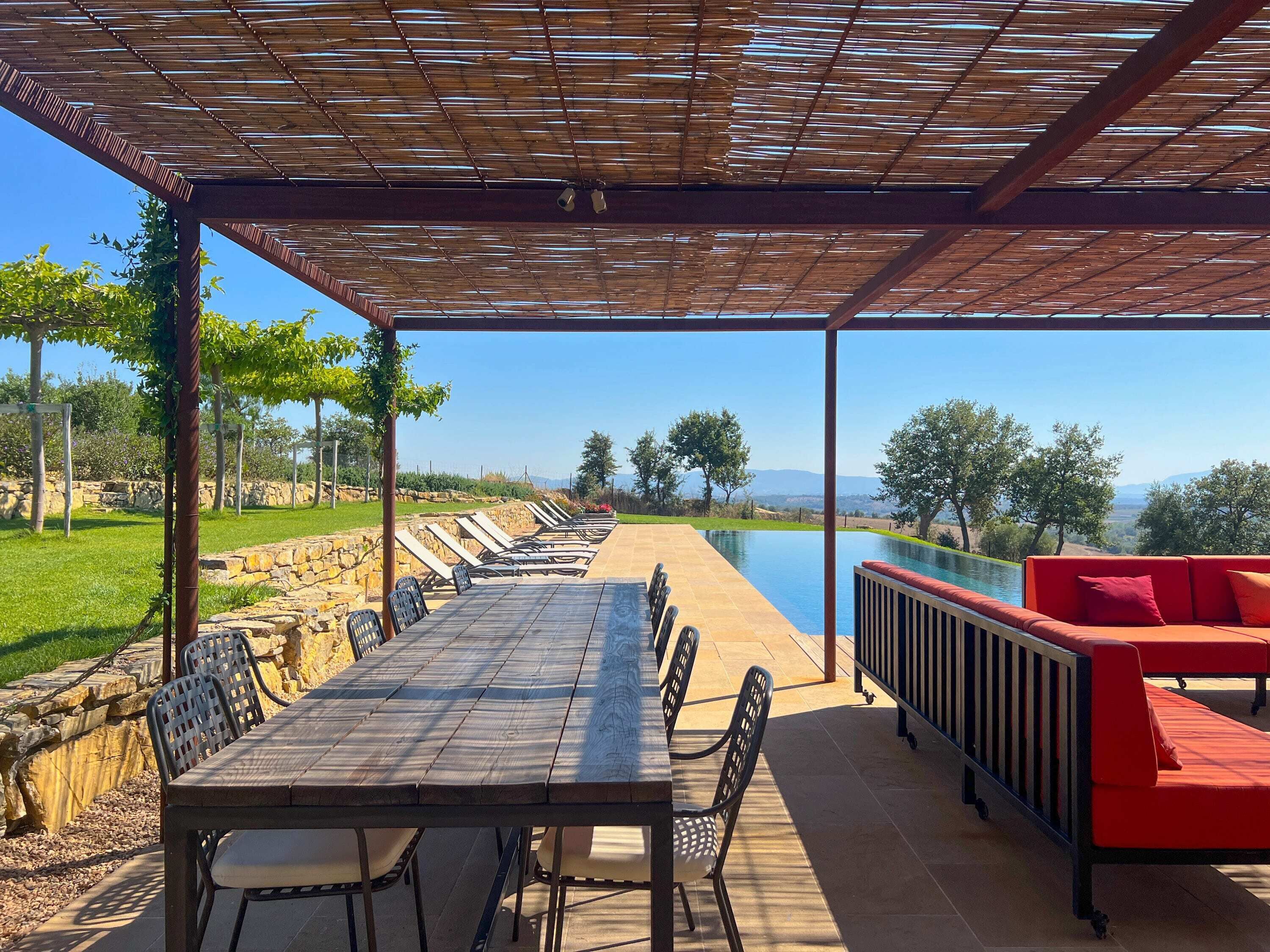 An outdoor patio with a long table and chairs, a red sofa, and a view of a pool and landscape.