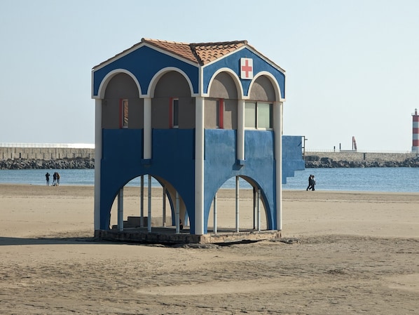 A blue and white building with arched windows and a red cross sign, located on a sandy beach with a rocky breakwater and a lighthouse in the background.