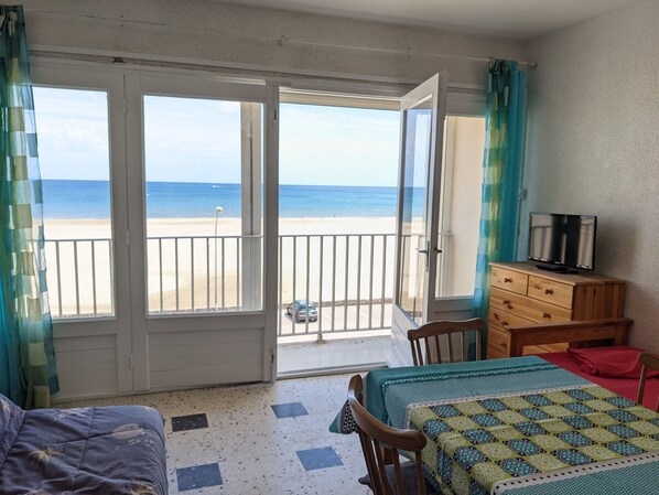 A room with a bed, a chair, a table, a TV, and a view of the beach and ocean.
