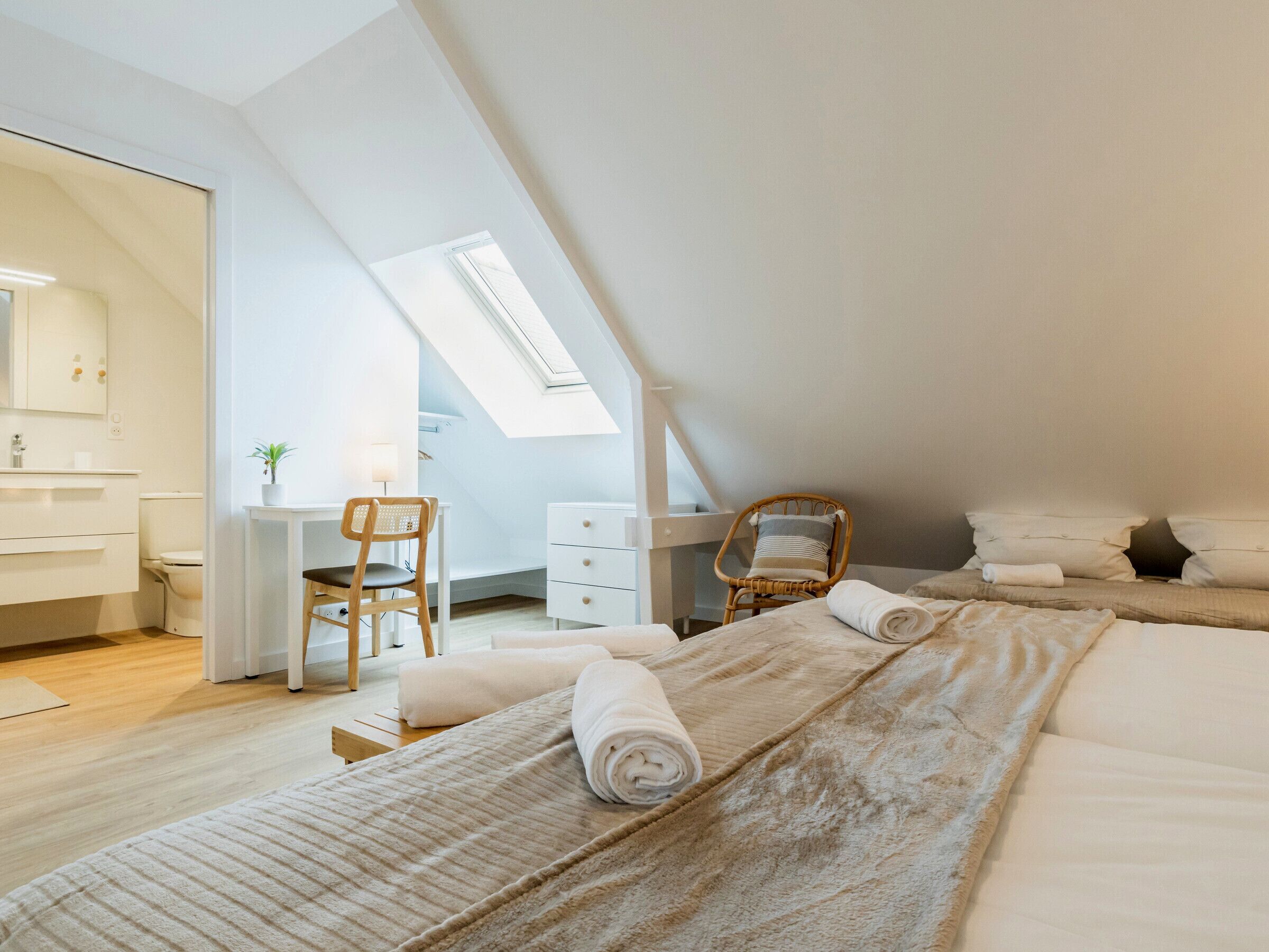 A modern, well-lit room with a skylight, a desk, a chair, and a bed with rolled towels.