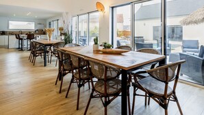 A spacious dining area with a long wooden table and wicker chairs, large windows offering views of the outdoors, and a modern kitchen in the background. - Luxurious Villa: Pool, Spa, Gym, Cinema, Near Beach, Sleeps 14 (La Turballe)