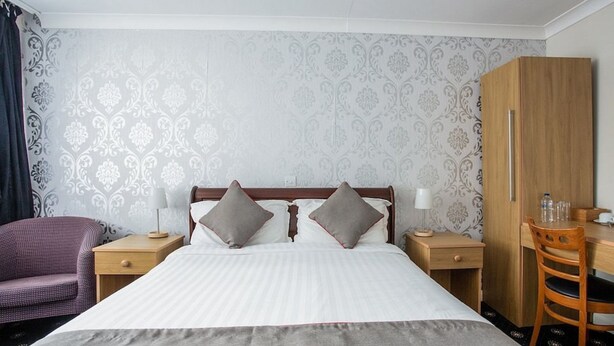 A hotel room with a large bed, two bedside tables, a wooden wardrobe, a desk with a chair, and a purple armchair. - Knowesgate Hotel (Newcastle-upon-Tyne)