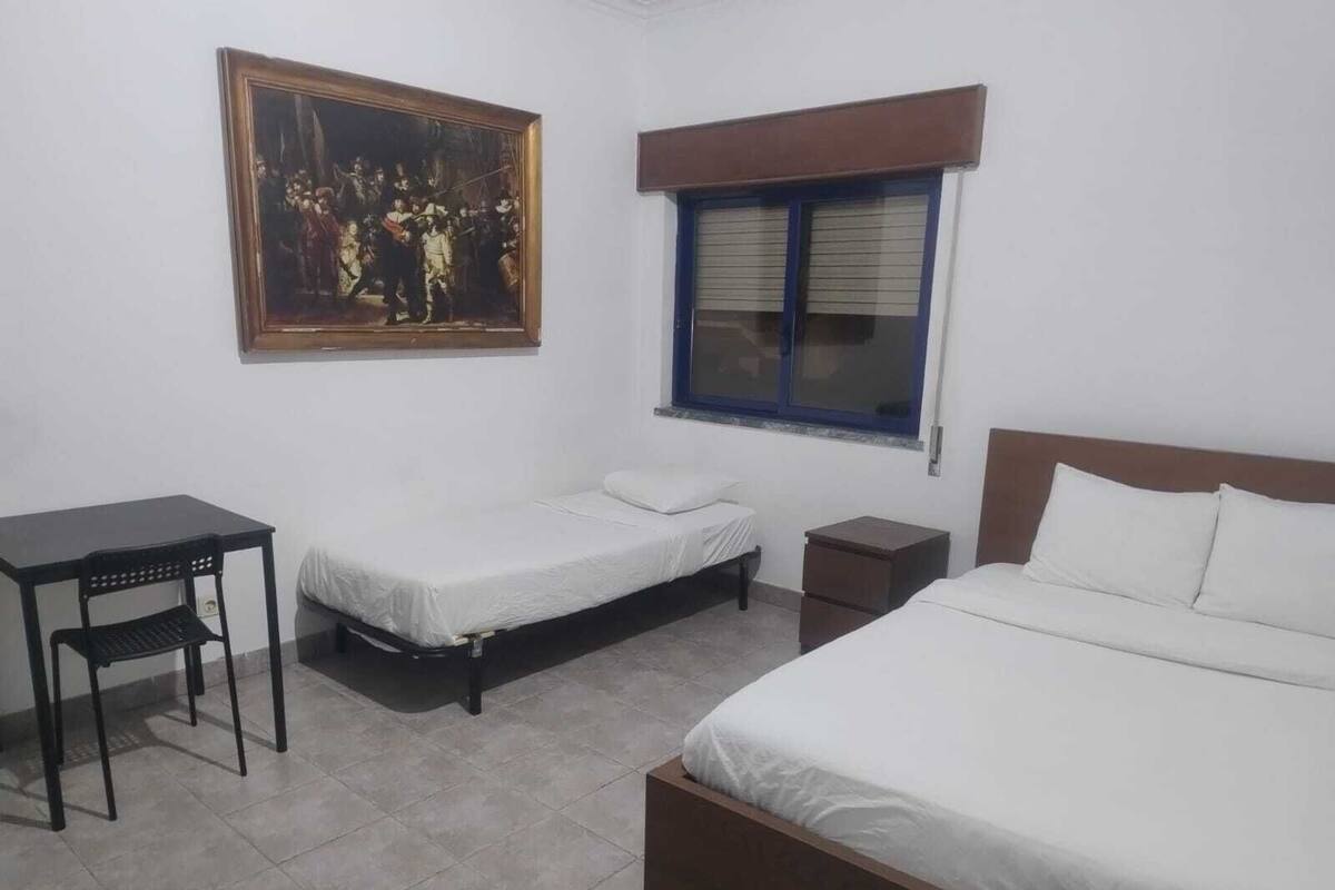 Ein Schlafzimmer mit zwei Einzelbetten, einem Schreibtisch, einem Stuhl, einem Nachttisch, einem Fenster mit Jalousien und einem gerahmten Bild an der Wand.