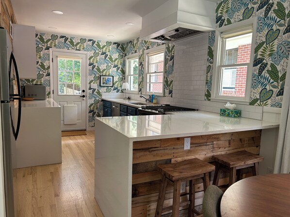 A modern kitchen with a large island, wooden bar stools, and a view of the outdoors through windows.