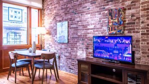 Una habitación con pared de ladrillo, una mesa de comedor de madera con sillas, un televisor sobre un mueble de madera y una ventana con un letrero.