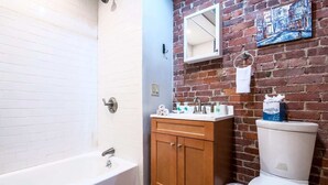 Un baño con una bañera blanca, tocador de madera y un inodoro frente a una pared de ladrillo.