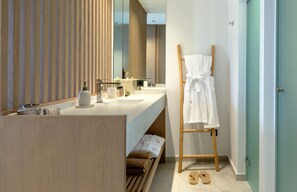 Une salle de bain moderne avec un meuble-lavabo en bois, un grand miroir, un porte-serviettes et une paire de chaussons posés sur le sol.