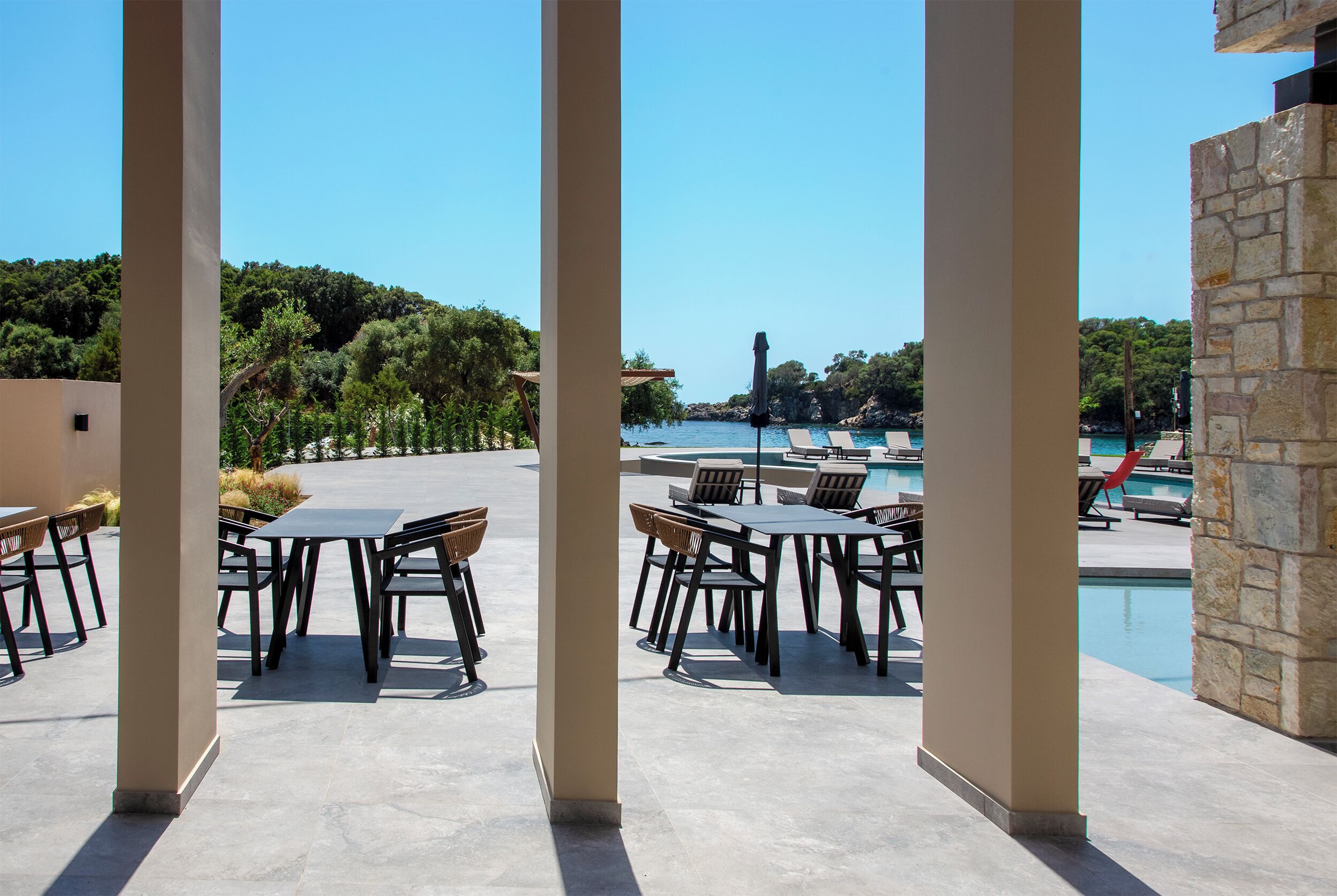 An outdoor pool area with lounge chairs and tables, surrounded by stone pillars and a view of the sea.