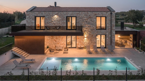 A two-story stone house with a swimming pool, outdoor seating, and a view of the surrounding landscape.