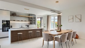 A modern kitchen with a central island, bar stools, a dining table with chairs, and a pendant light.