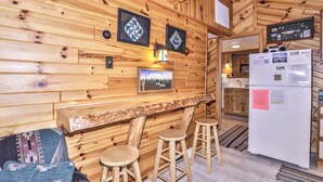 Interior em madeira com área de bar, banquetas, geladeira e cozinha visível ao fundo.