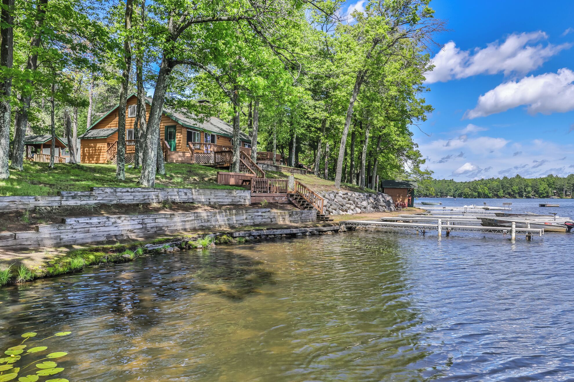 Une cabane en rondins avec un quai s’avançant dans un lac, entourée d’arbres et d’un mur de pierres.