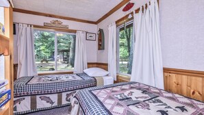 Quarto com duas camas, cabeceira de madeira, janela com cortinas brancas e vista para o exterior.