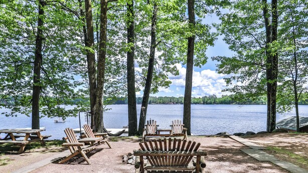 A lakeside campsite with wooden benches and picnic tables, surrounded by tall trees and a clear view of the lake. - Estrold's Resort- Driftwood Cabin (St.Germain)