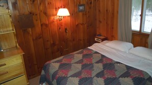 Une pièce aux murs lambrissés, contenant un lit, une commode, une lampe et une fenêtre.