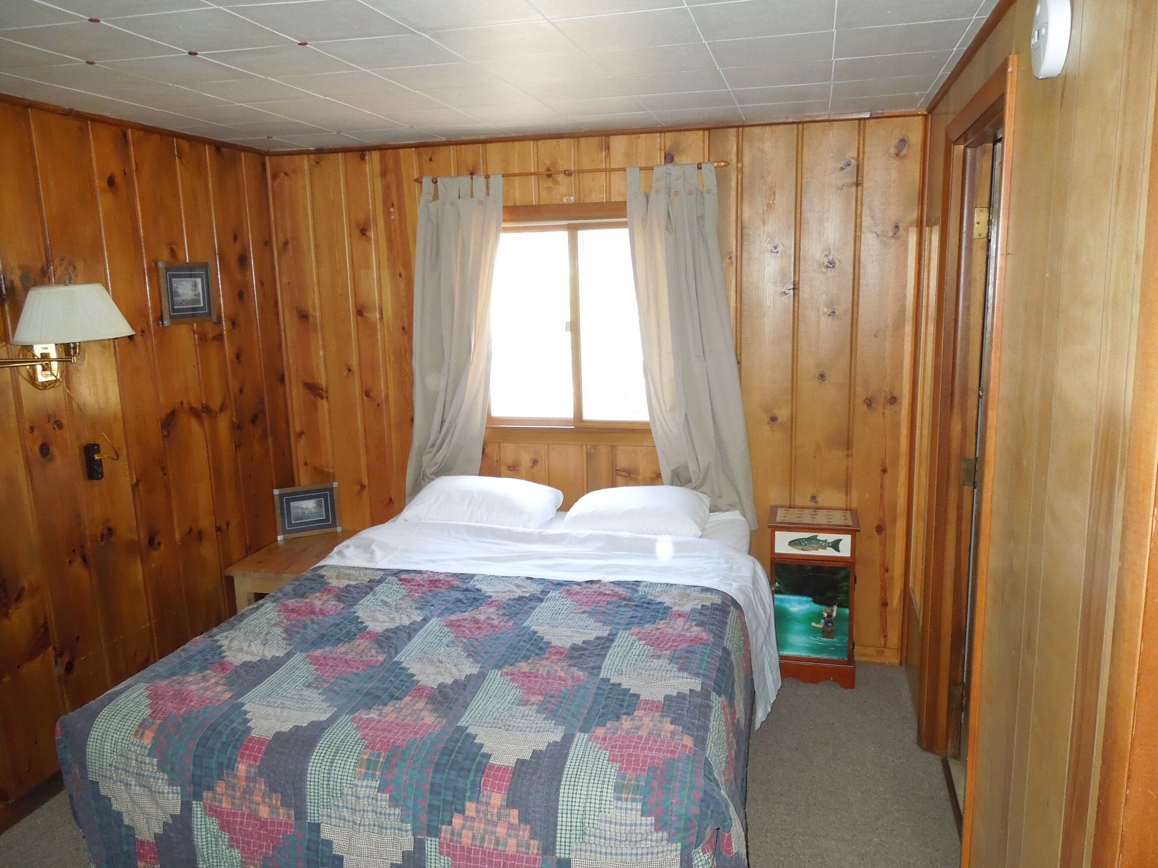A wooden-paneled room with a bed, a nightstand, a lamp, and a window with curtains.