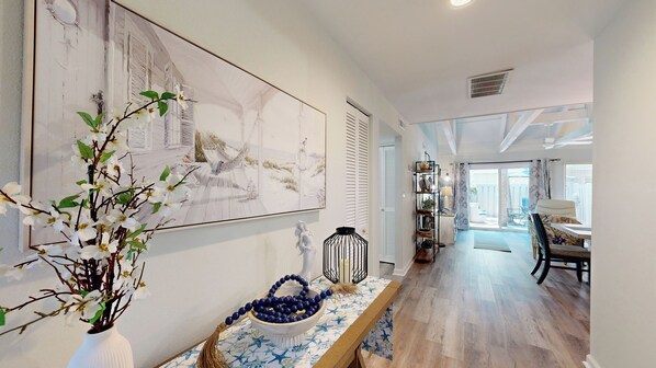 A spacious room with a large painting, a wooden bench with a blue and white patterned tablecloth, and a view of a balcony with outdoor furniture. - Recently Renovated Coastal Inspired Villa, Short Walk to the Beach! (Hilton Head)