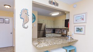 A kitchen with granite countertops, a refrigerator, and a coffee maker. There are two blue barstools and a white stool. - Oceanfront Building With Great Ocean and Bay Views (OCEAN OCEAN CITY)