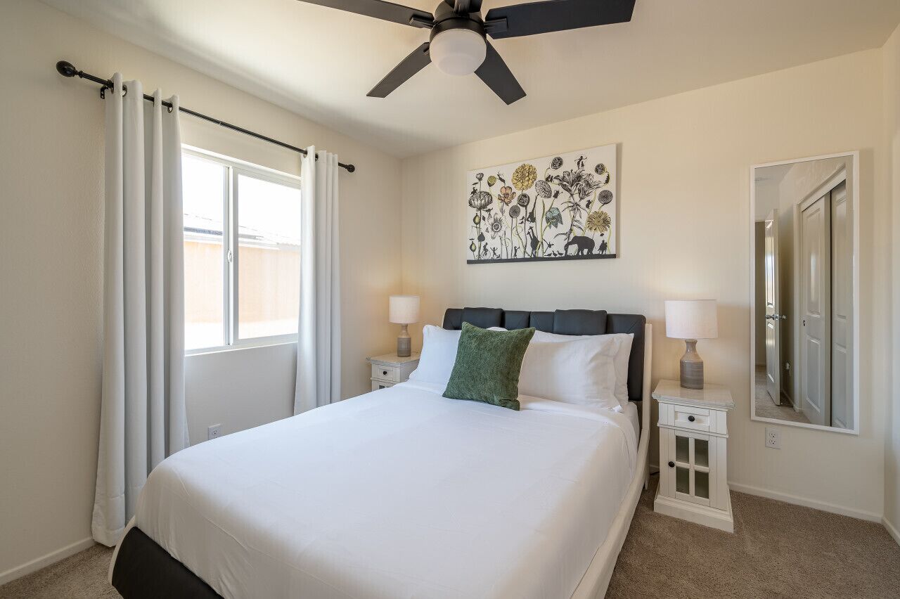 Une chambre à coucher comprenant un lit, deux tables de chevet avec des lampes, un ventilateur de plafond, une fenêtre avec des rideaux et une œuvre d’art accrochée au mur.