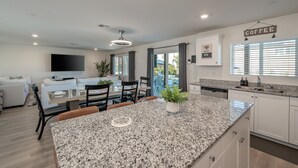 A modern kitchen with granite countertops, a dining area with a white table and chairs, and a living area with a sofa and TV. - Entertainer`s Paradise | Luxe Pool + Game Room (Coachella)