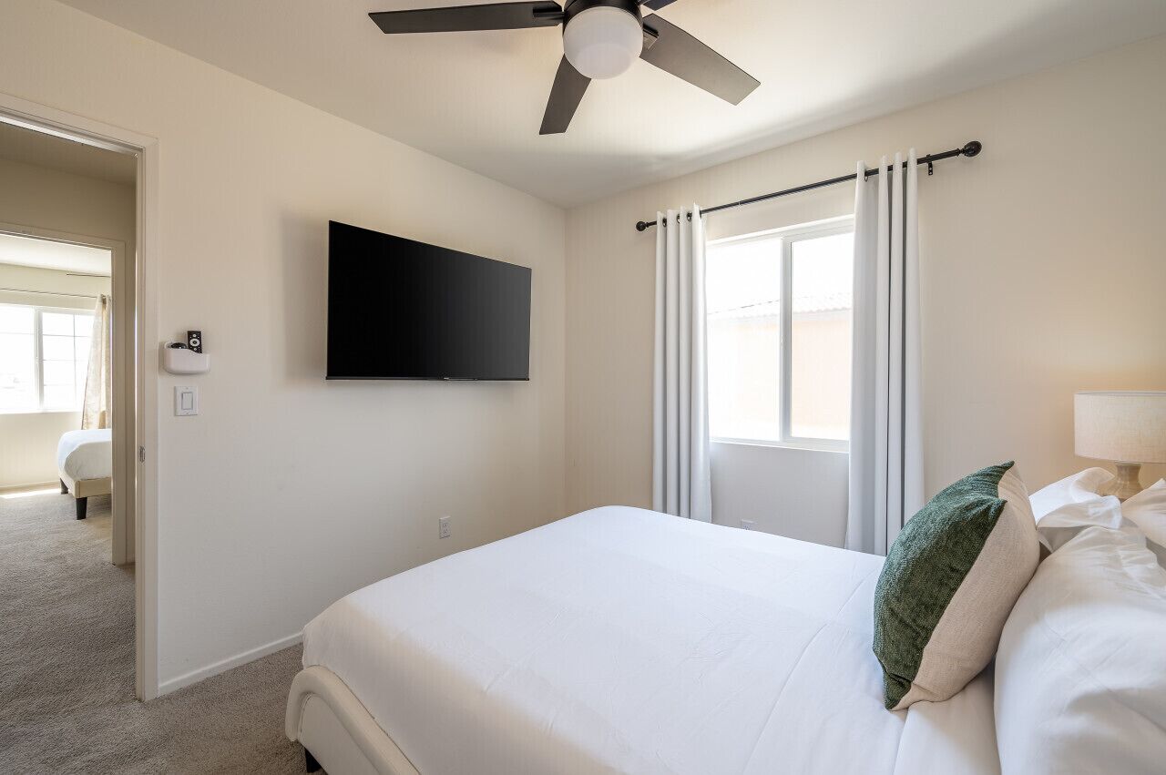 A hotel room with a bed, a ceiling fan, a TV mounted on the wall, and a window with curtains.