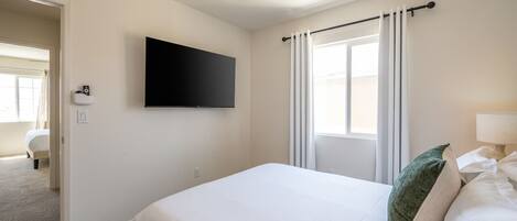A hotel room with a bed, a ceiling fan, a TV mounted on the wall, and a window with curtains.