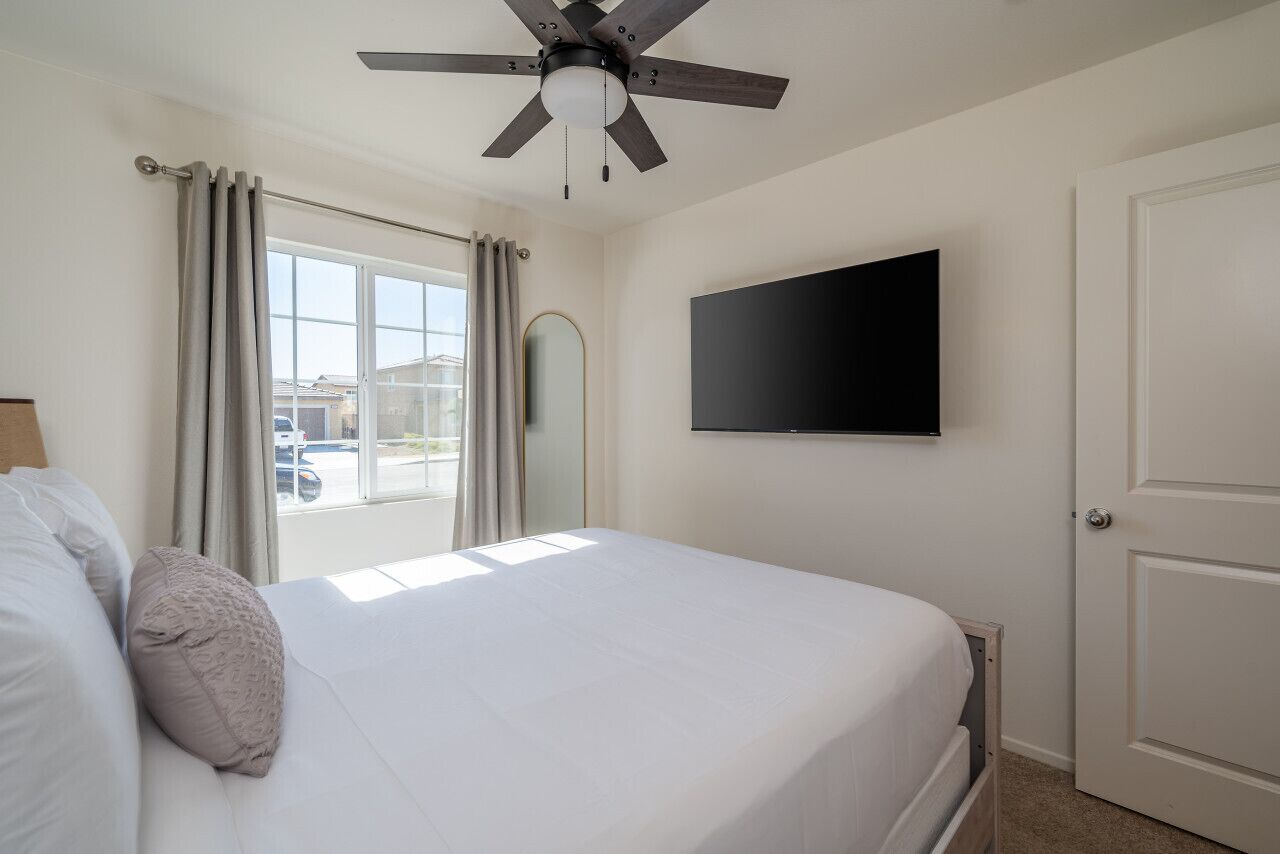 Un dormitorio con cama, ventilador de techo, televisor montado en la pared y una puerta.